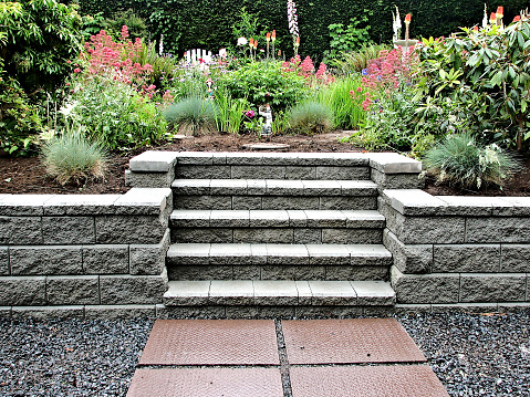 Block retaining wall with incorporated staircase into existing garden landscape consisting of perennial flowers and bushes with stepping stones leading up to the steps.