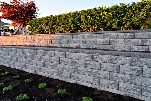 A close-up capture of a gray colored block stone retaining wall built as a two tier wall into an existing garden landscape viewed from the side in the early morning hours under sunny conditions.