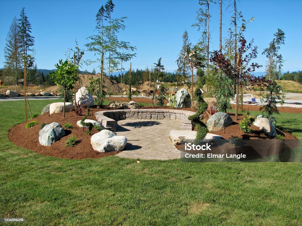 Horseshoe Shaped Deck New horseshoe shape paving stone deck with concrete block  retaining wall built on a mound of dirt filled with boulder - rocks, plants and trees surrounded by a large grass area in form of turf. Mulch Stock Photo
