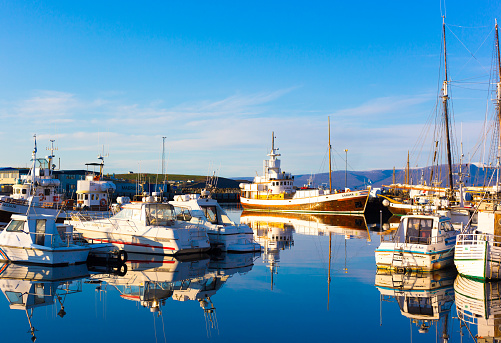 Húsavík, Iceland: Fishing trawlers and an antique sailboat in Húsavík’s sunlit harbor in early morning in September, with snowcapped mountains in the background. Húsavík is a fishing town as well as whale-watching center in North Iceland.