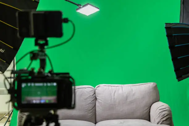 a gray couch stands in front of a green screen. Background interchangeable. in the foreground you can see a camera and softboxes. Interviewsetting with colored background.
