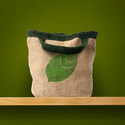 Burlap tote bag printed with green leaf on a shelf against green wall with copy space. 
Concept of environmental and recycling.