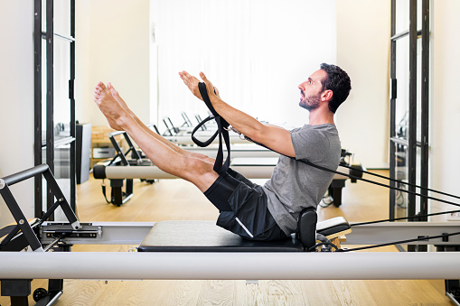 Fit muscular man doing a teaser pilates exercise on a reformer in a gym to strengthen his core and abdominal muscles in a side view with copyspace