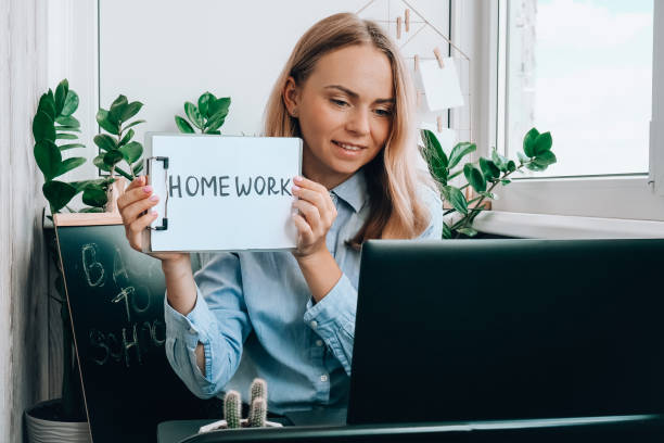 istock Young female teacher sitting at desk and using laptop showing HOMEWORK text at home. e Education Back to school. Student learning online class. Distance learning. Exam preparation 1336617667