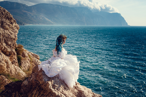 Young woman standing on cliff's edge and looking into a wide sea view. Wind Long Blue hair hairstyle.