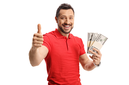 Happy young man in a red t-shirt holding money and showing thumbs up isolated on white background