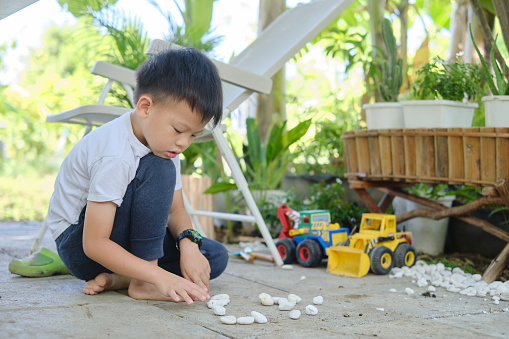 Cute Asian young kindergarten boy playing with pebbles and toy construction machinery, Happy smiling kid playing alone at home backyard outdoor on nature, Montessori education, Creative play for kids