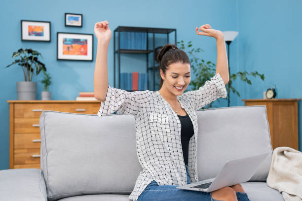 istock A beautiful brunette sits on a couch at home with a laptop on lap, video chatting, webcam, discussing the internet with a friend, explaining, telling stories, gesturing hands up, smiling, joking 1336621794