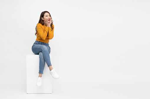 Portrait of excited screaming young asian woman sitting on white box isolated over white background, Wow and surprised concept