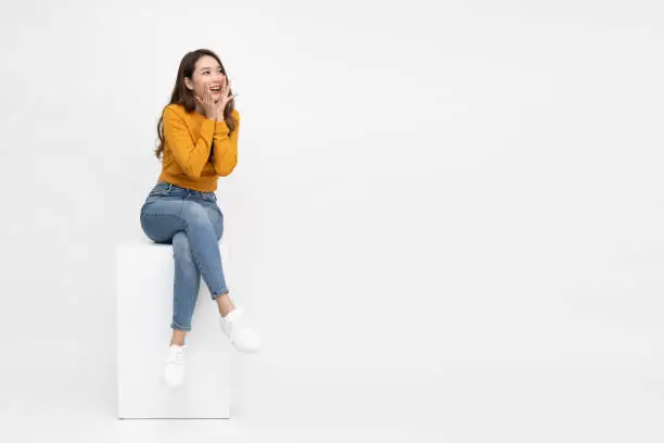 Photo of Portrait of excited screaming young asian woman sitting on white box isolated over white background, Wow and surprised concept