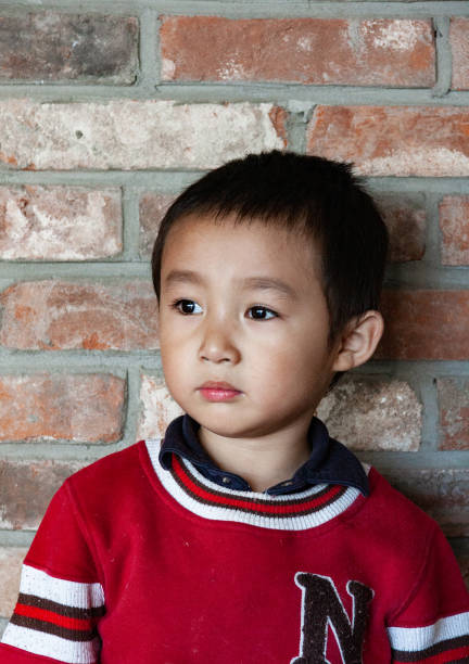 A portrait of a handsome Asian boy wearing a sweater standing against a wall in natural light.