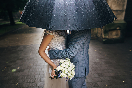 Stylish bride with bouquet and groom kissing under umbrella on background of old church in rain. Provence wedding. Beautiful wedding couple embracing under black umbrella in rainy street