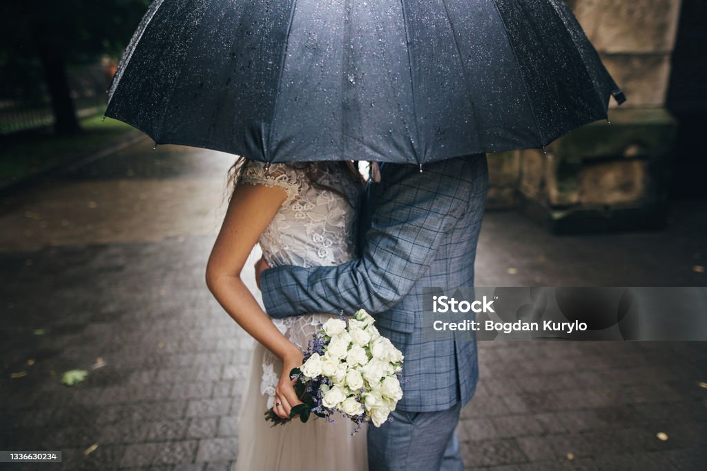 Stylish bride with bouquet and groom kissing under umbrella on background of old church in rain. Provence wedding. Beautiful wedding couple embracing under black umbrella in rainy street Rain Stock Photo