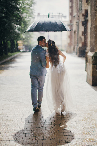 Rain drops on background of blurred stylish bride and groom walking under umbrella and kissing at old church in rain. Provence wedding. Beautiful wedding couple embracing in sunny rain
