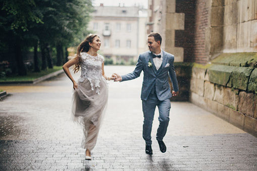 Blurred image of beautiful emotional wedding couple smiling in rain in european city. Provence wedding. Stylish happy bride and groom running on background of old church in rainy street.