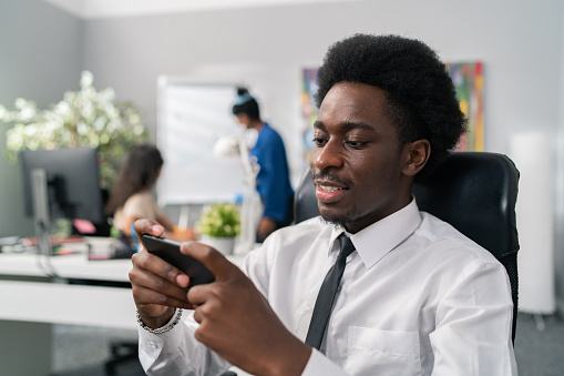 Elegant handsome executive in afro and dark skin sits comfortably leaning on a chair dressed in white shirt and tie, spends his free time in the office playing games on phone