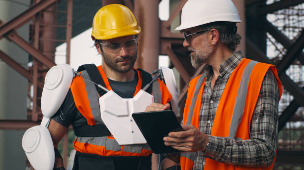 Male contractors discussing plan on tablet Zoom out view of mature foreman in sunglasses using tablet and discussing work strategy with adult employee in exoskeleton on construction site powered exoskeleton photos stock pictures, royalty-free photos & images