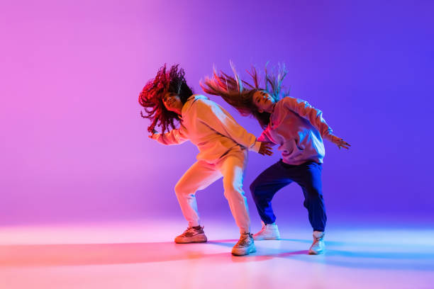 Two beautiful active girls dancing on gradient pink purple neon studio background Active lifestyle. Two beautiful hip-hop dancers in motion on gradient pink purple neon background. Sport achievement, expression. Concept of dance, youth, hobby, dynamics, movement, action, ad disco dancing photos stock pictures, royalty-free photos & images