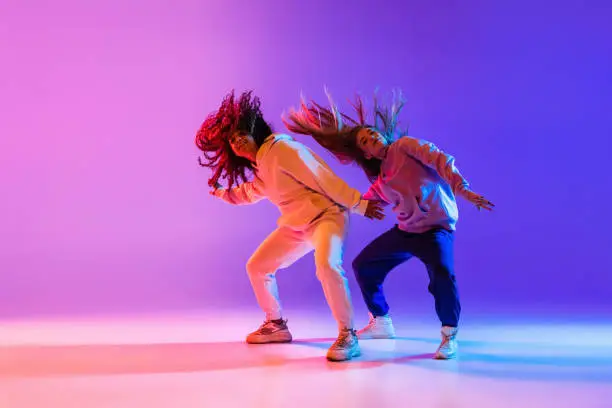 Photo of Two beautiful active girls dancing on gradient pink purple neon studio background