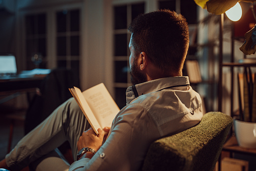 One man, charming male sitting on a chair and reading a book alone. at home at night.