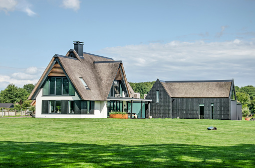 Heino, The Netherlands, August 7, 2021: modern villa with traditional materials like stucco, wood, and a thatched roof on a lawn kept short by a robotic lawn mower