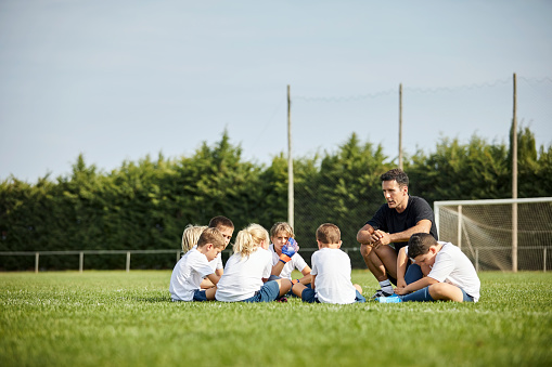 Soccer coach talking to children. Girls and boys are sitting on grass. They are in uniform.