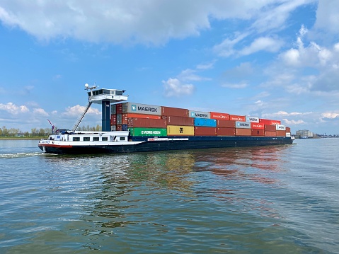 Inland barge freight shipping transport sailing pass Dordrecht in Holland in the Netherlands on a calm river water of the Maas. Container ship transporting goods in colourful metal containers. Scenic Dutch summer sky urban landscape by the Groothoofd in Dordt. Concept and copy space.