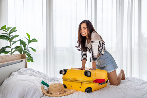 Happy young Asian woman packing and trying to close luggage, smiling. Girl preparing suitcase for summer vacation travel trip, looking at camera.