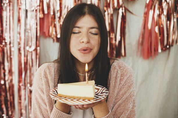 スタイリッシュな幸せな女性は、誕生日ケーキの一部に燃えるろうそくを吹いて、部屋にモダンなローズゴールドのタッセルガーランドの背景に願いを作ります。自宅で誕生日を祝う - cake birthday candle blowing ストックフォトと画像