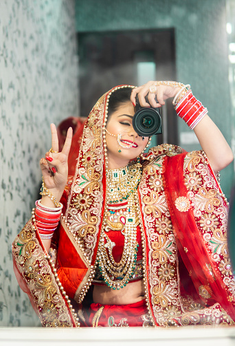 Side view portrait of beautiful, happy traditional Indian bridal in red lehenga and jewelry. She is taking selfie in the mirror with camera and showing victory finger.