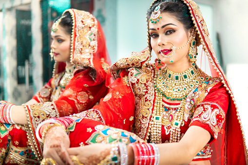 Side view portrait of beautiful traditional Indian bridal in red lehenga and jewelry. She is sitting near mirror and looking at the camera with.