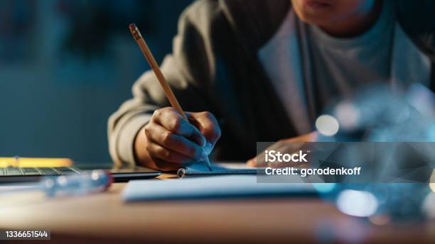 Close Up Of A Teenage Multiethnic Black Girl Writing Down Homework In A Notebook With A Pencil Using Laptop Computer In A Dark Cozy Room At Home Shes Browsing Educational School Research Online Stock Photo - Download Image Now