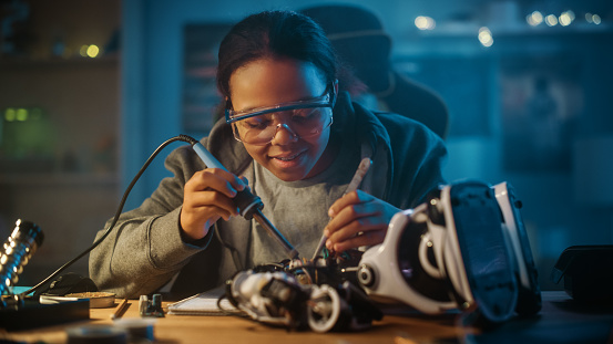 Una joven adolescente multiétnica está estudiando electrónica y soldadura de cables y placas de circuitos en su proyecto de robótica De Pasatiempo Científico. La chica está trabajando en un robot en su habitación. Concepto de educación. photo