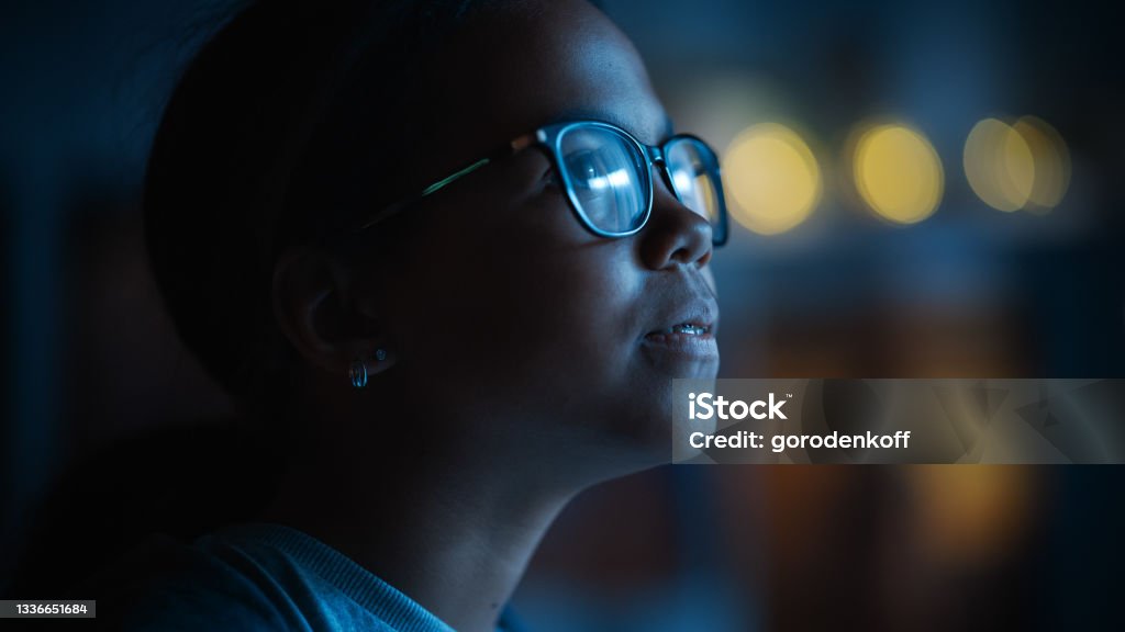 Portrait of a Teenage Multiethnic Black Girl Looking Out of the Window in Excitement. Surprised Young Female Watching the Night Sky from Her Home. She Wears Glasses and Dental Braces. Dark Stock Photo