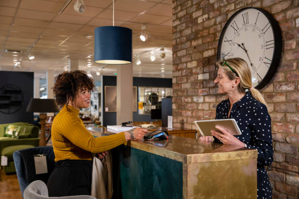 Making a Furniture Purchase A mid adult woman making a contactless payment in a furniture shop. She is tapping her card on the card reader while looking at the store employee who is standing at the cash register desk holding a digital tablet. point of sale tablet stock pictures, royalty-free photos & images