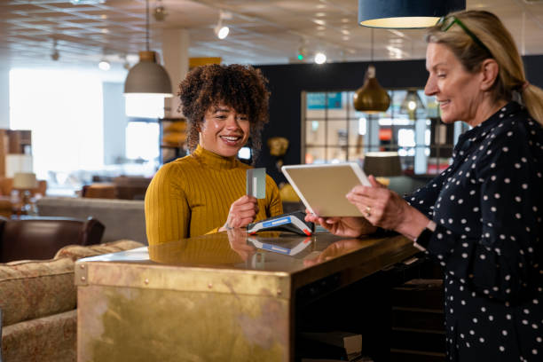Giving Her a Price Breakdown A mid adult woman making a contactless payment in a furniture shop. She is holding her credit card while smiling and looking at a digital tablet that the store employee is showing to her. point of sale tablet stock pictures, royalty-free photos & images