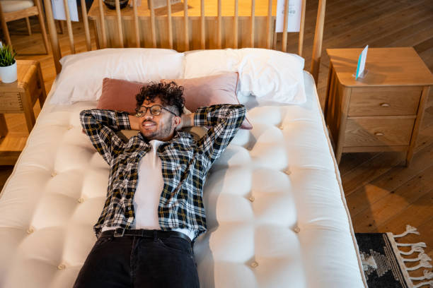 Testing Out Beds A mid adult man lying on a bed in a furniture store, he is testing it out and considering to buy it. He has his arms behind his head and is looking away from the camera. mattress stock pictures, royalty-free photos & images