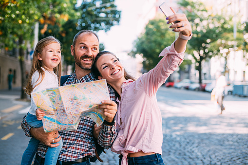 Shot of a father, mother and their little daughter exploring in a foreign city. Father checking city map while mother making selfie with smart phone.