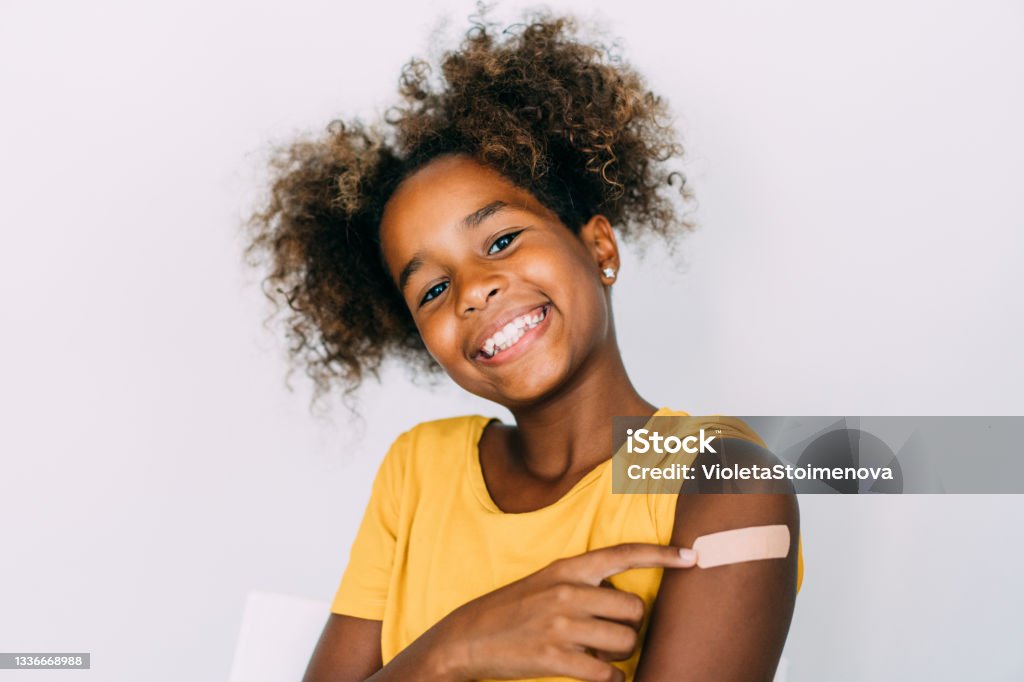 Little girl showing her arm after getting vaccinated. Cute smiling girl pointing at her arm with a bandage after receiving COVID-19 vaccine. Shot of little african-american girl showing her shoulder after getting coronavirus vaccine. Vaccination Stock Photo