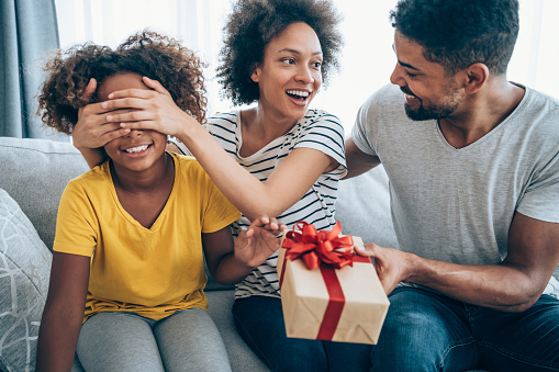 Shot of a loving mother and father giving their daughter a gift. Parents surprise their little girl with present while she is sitting on the sofa in the living room at home.