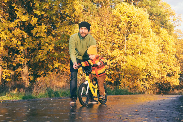 father teaches his little kid to ride bike in autumn park. child practicing cycling. happy family moments. time together dad and son. candid lifestyle photo. - candid people casual bicycle imagens e fotografias de stock