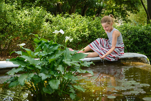 Teen girl is sitting on the shore of small decorative garden pond and playing with water