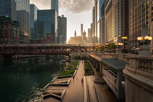 Hermosa mañana en el centro de Chicago a lo largo del río mientras la gente trota por el camino de abajo y el tren cruza un puente mientras el sol proyecta luz amarilla en la escena desde detrás de los edificios de gran altura más allá. photo