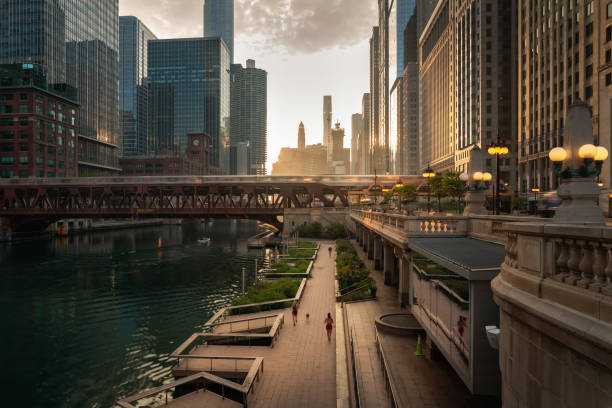 bella mattinata nel centro di chicago lungo il fiume mentre le persone fanno jogging sul sentiero sottostante e il treno attraversa un ponte mentre il sole proietta luce gialla sulla scena da dietro i grattacieli oltre. - chicago skyline illinois downtown district foto e immagini stock