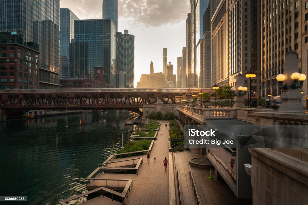 Beautiful downtown Chicago morning along the river as people jog on the path below and train crosses a bridge as the sun casts yellow light into the scene from behind the high-rise buildings beyond. Chicago - Illinois Stock Photo