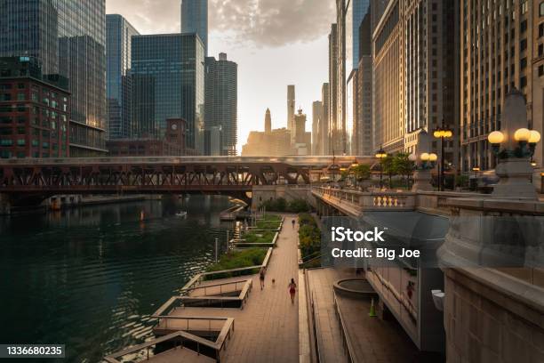 Beautiful Downtown Chicago Morning Along The River As People Jog On The Path Below And Train Crosses A Bridge As The Sun Casts Yellow Light Into The Scene From Behind The Highrise Buildings Beyond Stock Photo - Download Image Now