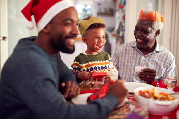 une famille de plusieurs générations en chapeaux de papier lisant des blagues de craquelins de noël avant de manger ensemble - christmas cracker photos et images de collection