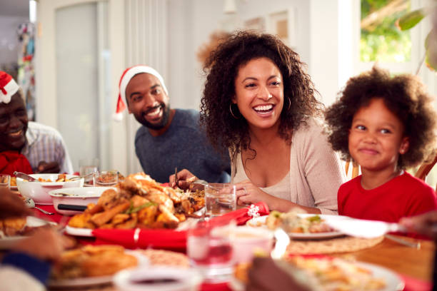 familia de varias generaciones con sombreros de papel disfrutando juntos comiendo comida de navidad en casa - los niños y los padres brindis bebidas mientras almuerza fotografías e imágenes de stock