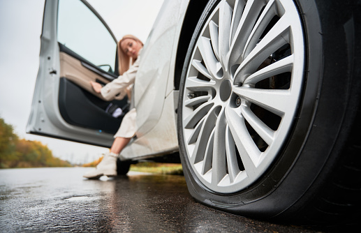 Close up view of rear semi-lowered wheel of white automobile parked on wet road. Blonde female driver stopped her car on edge of road and looking at punctured tire of back wheel.