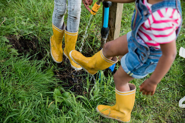 A high angle over the shoulder view of two sisters cleaning their wellies on an outdoor tap next to their allotment near their home in the North East of England. They are wearing wellington boots and are their faces also cant be seen in the shot.